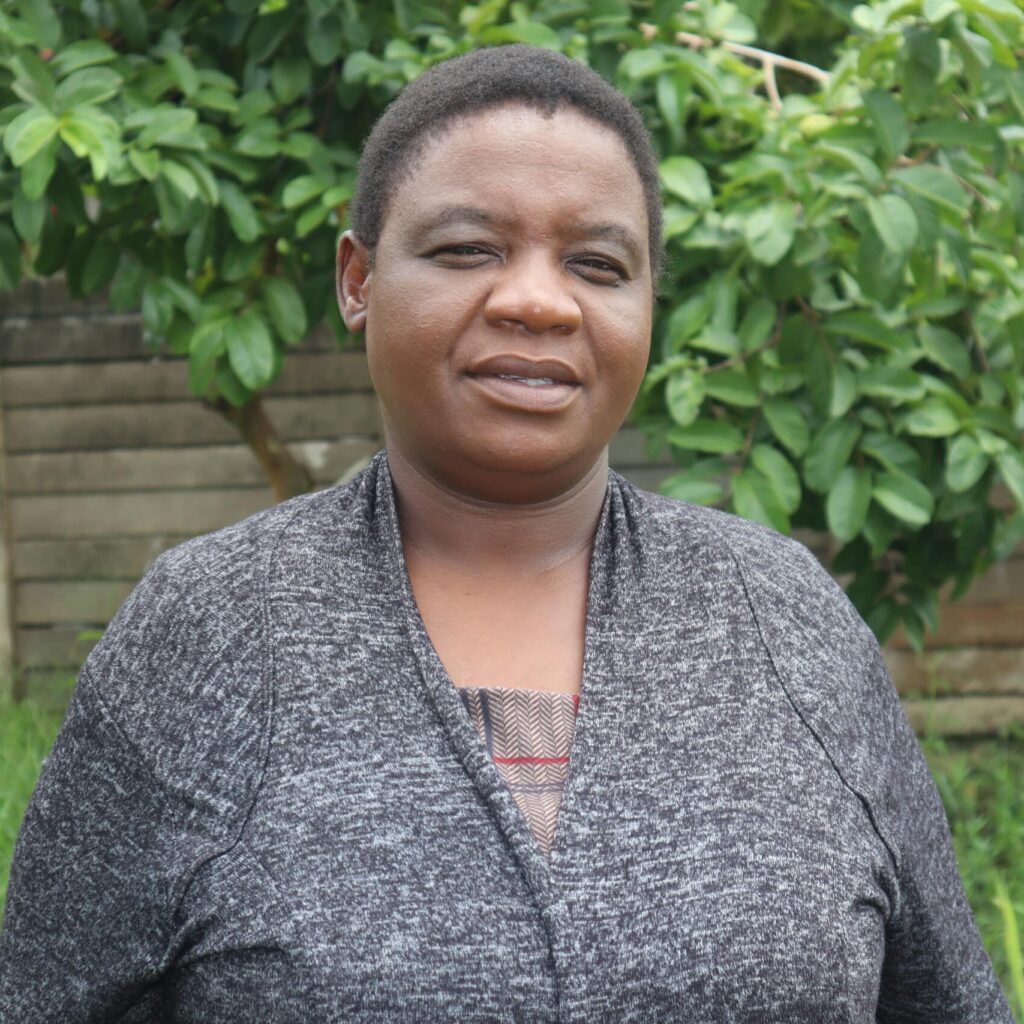 A photo of a dark skinned Zimbabwean woman wearing a dark grey cardigan against a background of green foliage.